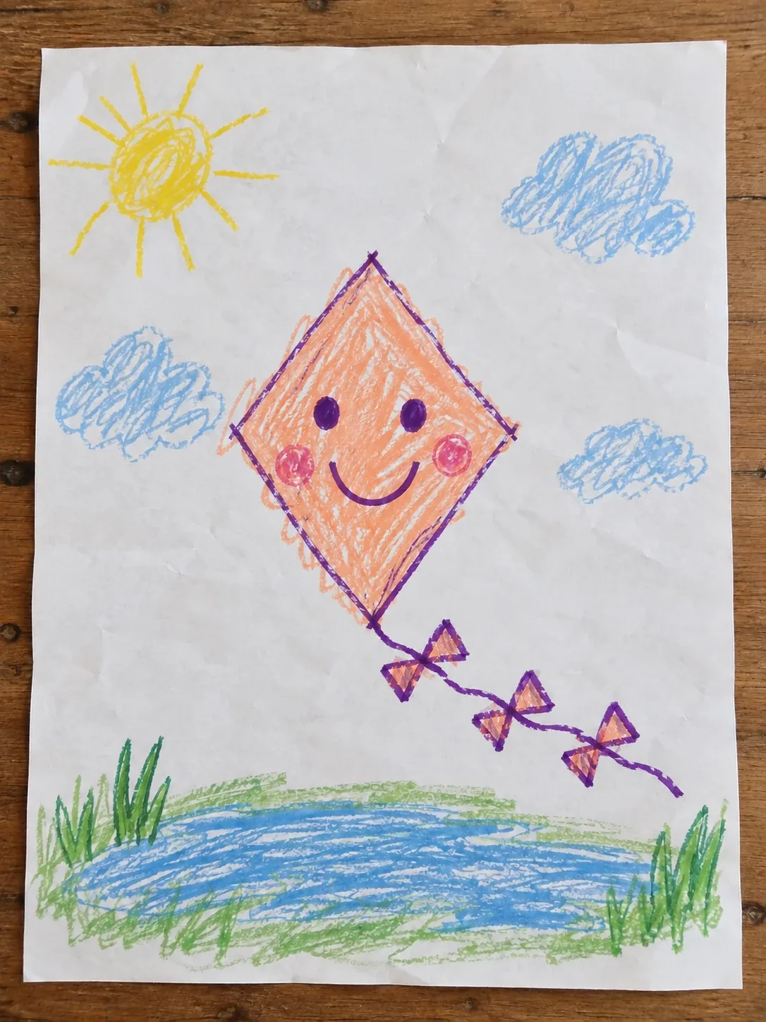 Dibujo infantil a crayón de una cometa sonriente con tres lazos sobre un estanque